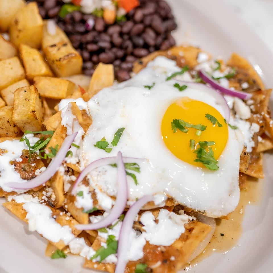 Chilaquiles with a fried egg, sour cream, red onions, cilantro, and a side of potatoes and black beans.