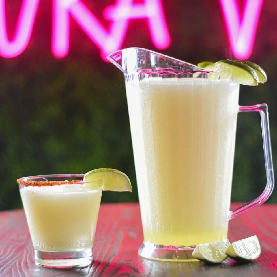 A margarita glass and pitcher with lime slices, against a neon pink sign.