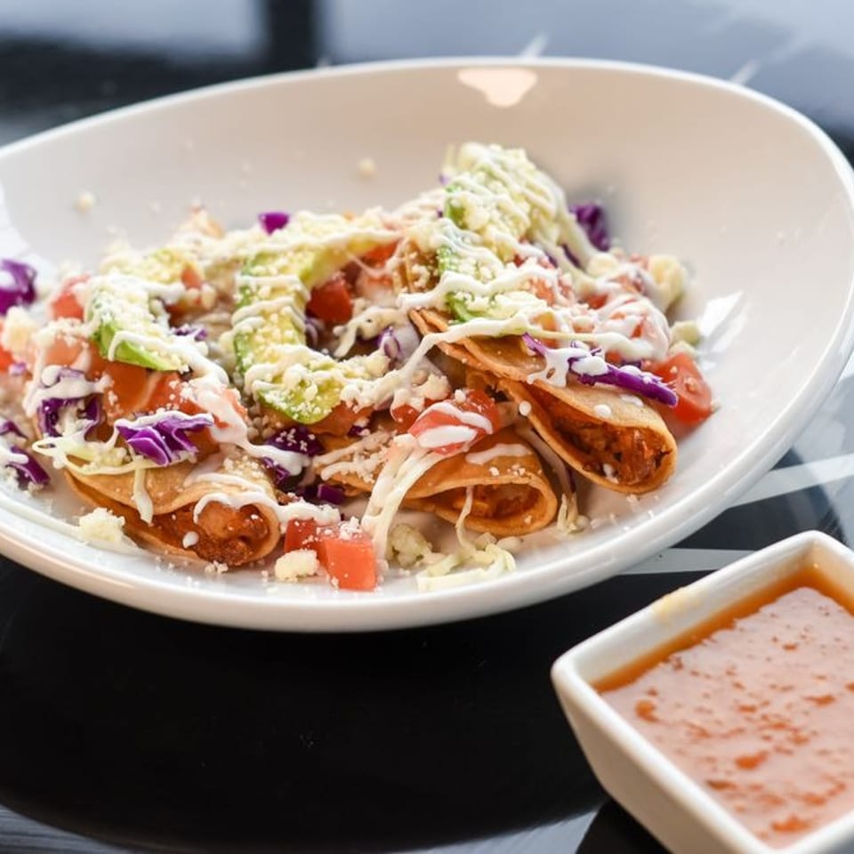 Crispy taquitos topped with avocado, tomatoes, shredded cabbage, cheese, and crema, with a side of salsa.