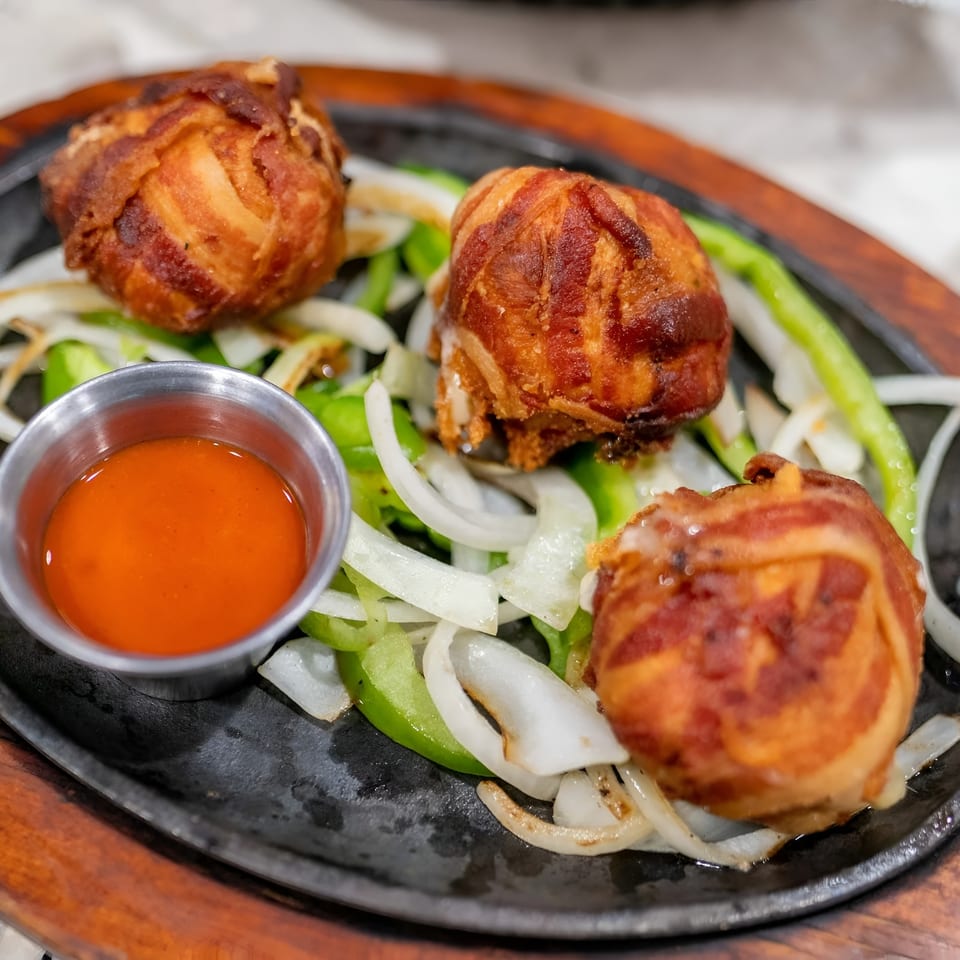 Three bacon-wrapped appetizers with onions and peppers on a sizzling platter with dipping sauce.