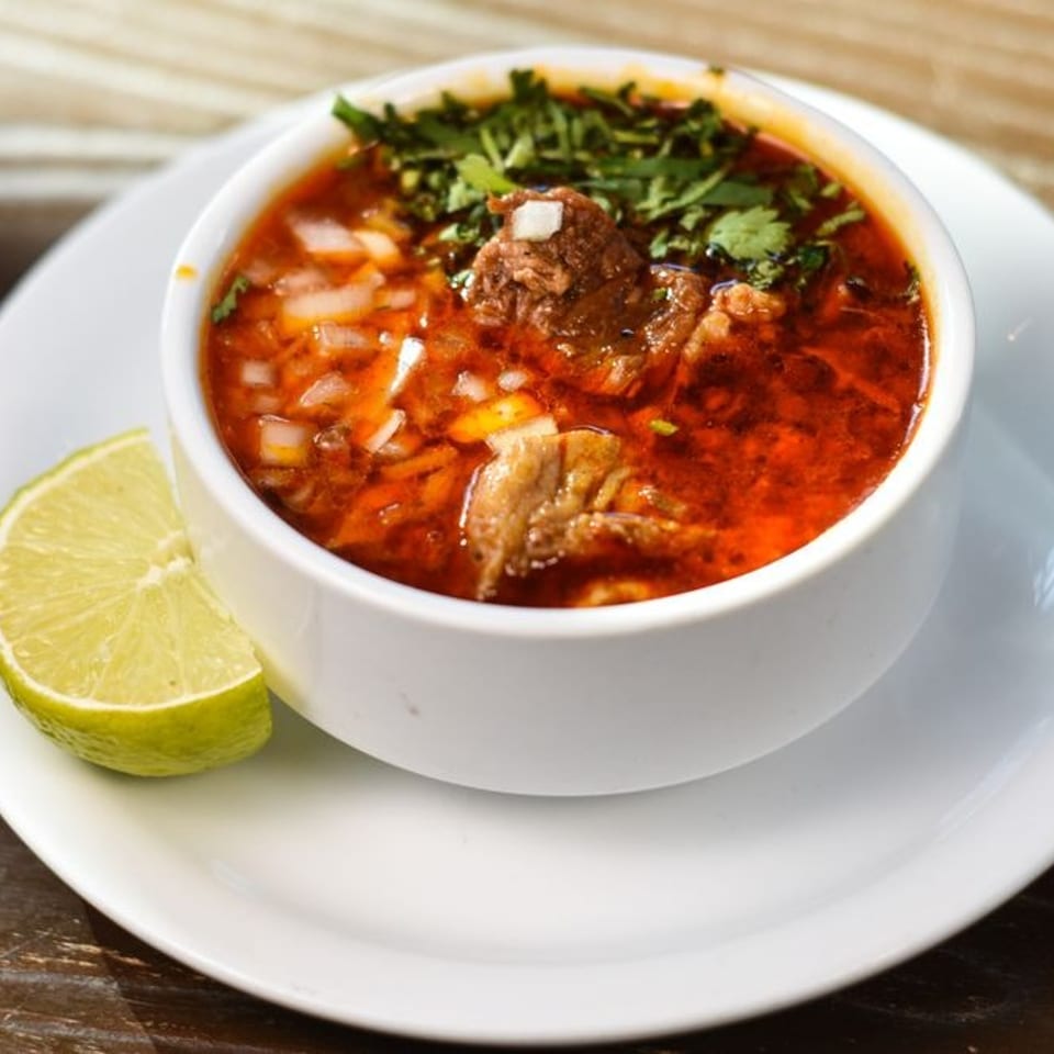 Hearty red soup with meat, onions, and cilantro in a white bowl with a lime wedge.