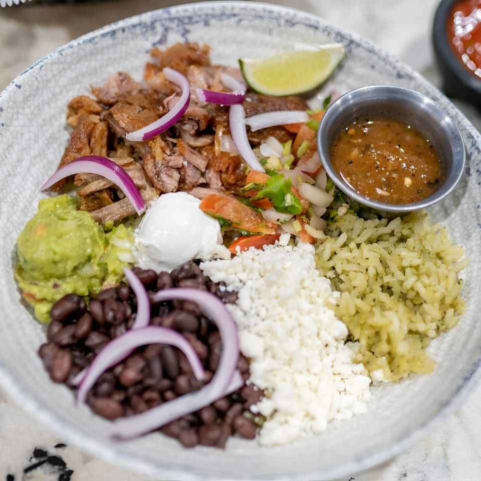 A colorful bowl of carnitas, rice, black beans, guacamole, cheese, and salsa on a marble counter.