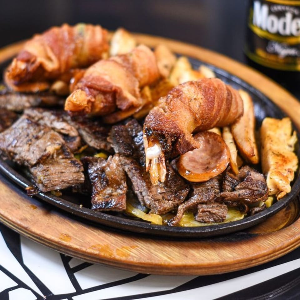 Sizzling platter of steak, bacon-wrapped shrimp, sausage, and fries, with a Modelo beer in the background.