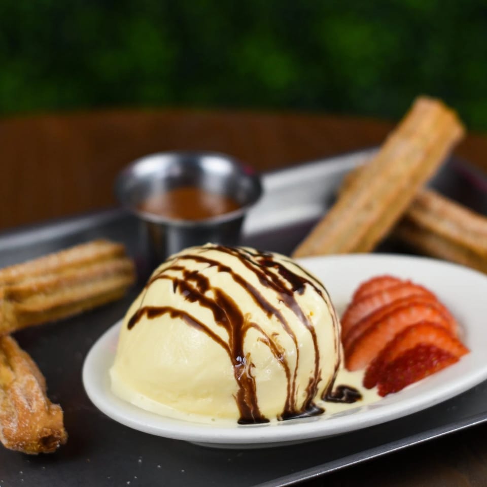 Churros with vanilla ice cream drizzled with chocolate, sliced strawberries, and a side of caramel.