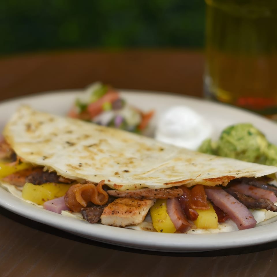 A quesadilla filled with chicken, ham, pineapple, and bacon, served with guacamole, sour cream, and salsa.