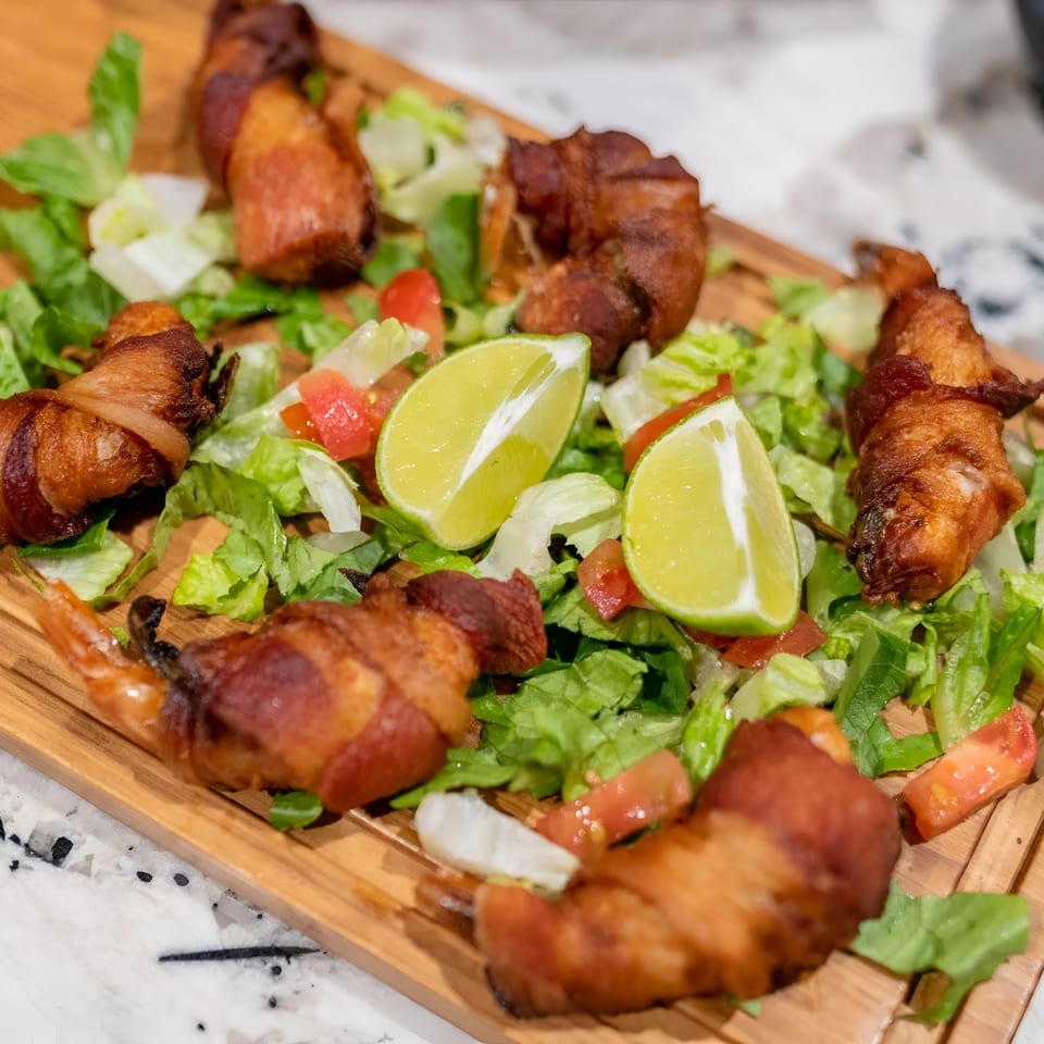 Bacon-wrapped shrimp on a bed of lettuce and diced tomatoes with lime wedges and a side of sauce.
