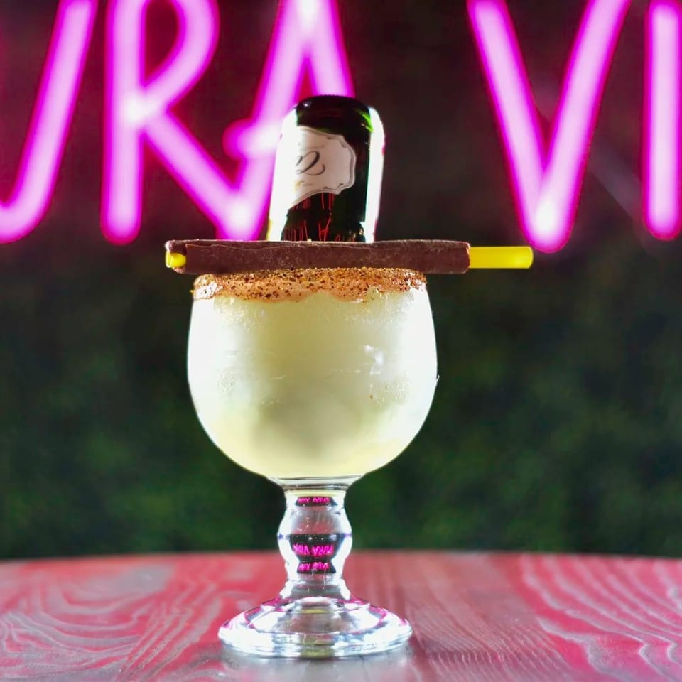 Frozen margarita with a mini beer bottle, chocolate stick, and yellow straw against a neon sign.