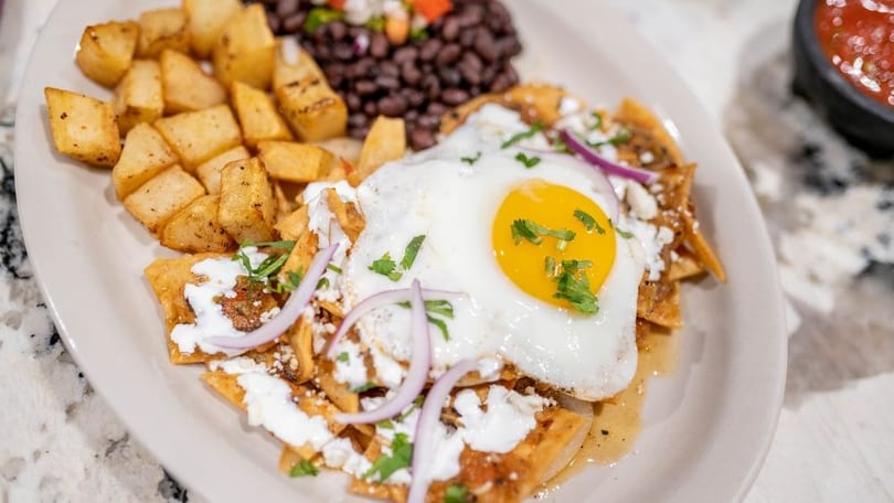 Chilaquiles with a fried egg, black beans, and roasted potatoes on a white plate.